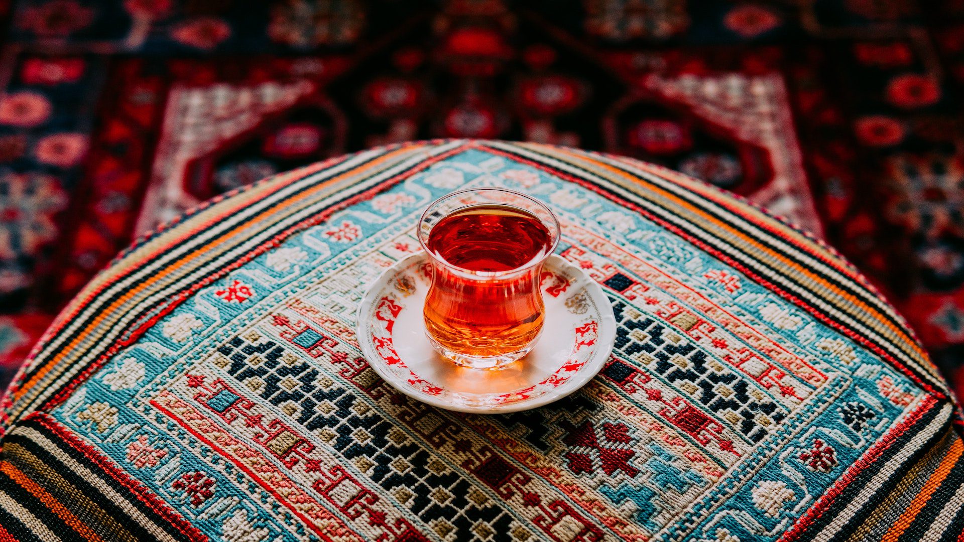 A glass of tea on plate on top of a colorful, patterned ottoman and rug.