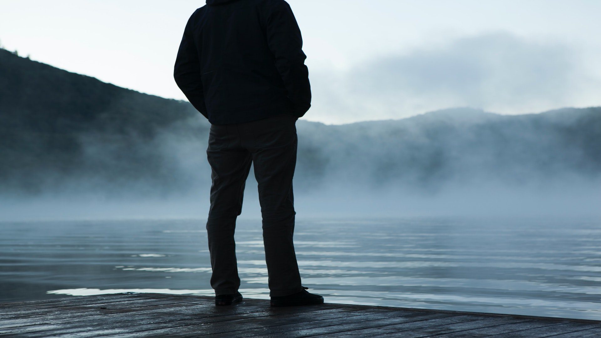 The back of a man standing on a dock near a lake covered with fog.