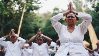 A woman in a white shirt with people behind her kneeling with their hands folded above their heads.