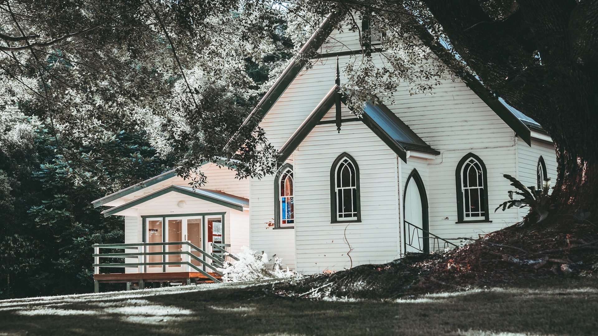 A white church with a black roof and stained glass windows with trees and grass surrounding it.