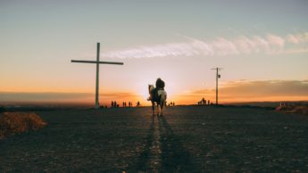 Person riding a horse on a field with a cross during a sunset.