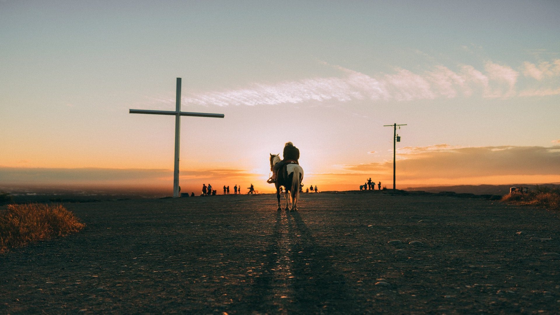 Person riding a horse on a field with a cross during a sunset.