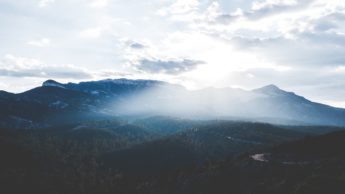 Sun shining through clouds over tree covered mountains.