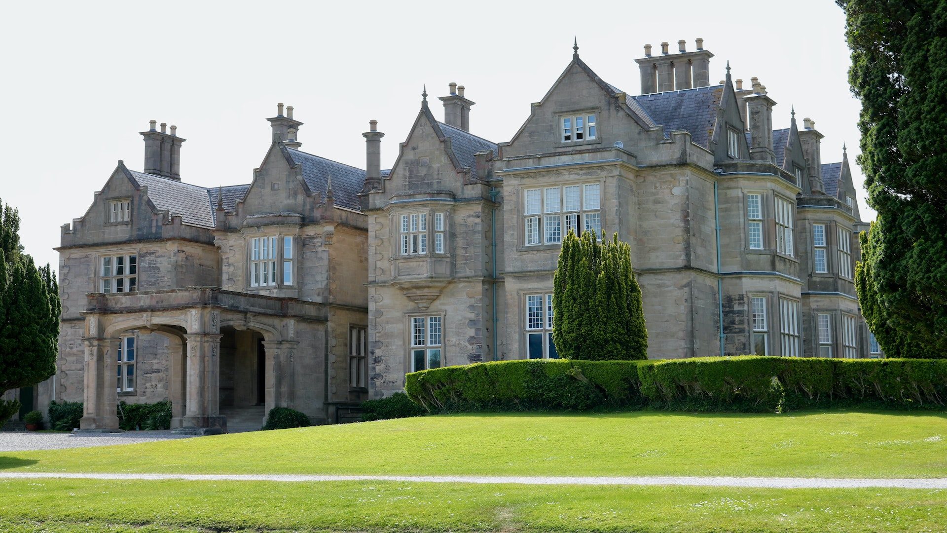 A brown, stone mansion surrounded by green grass, shrubs, and trees.