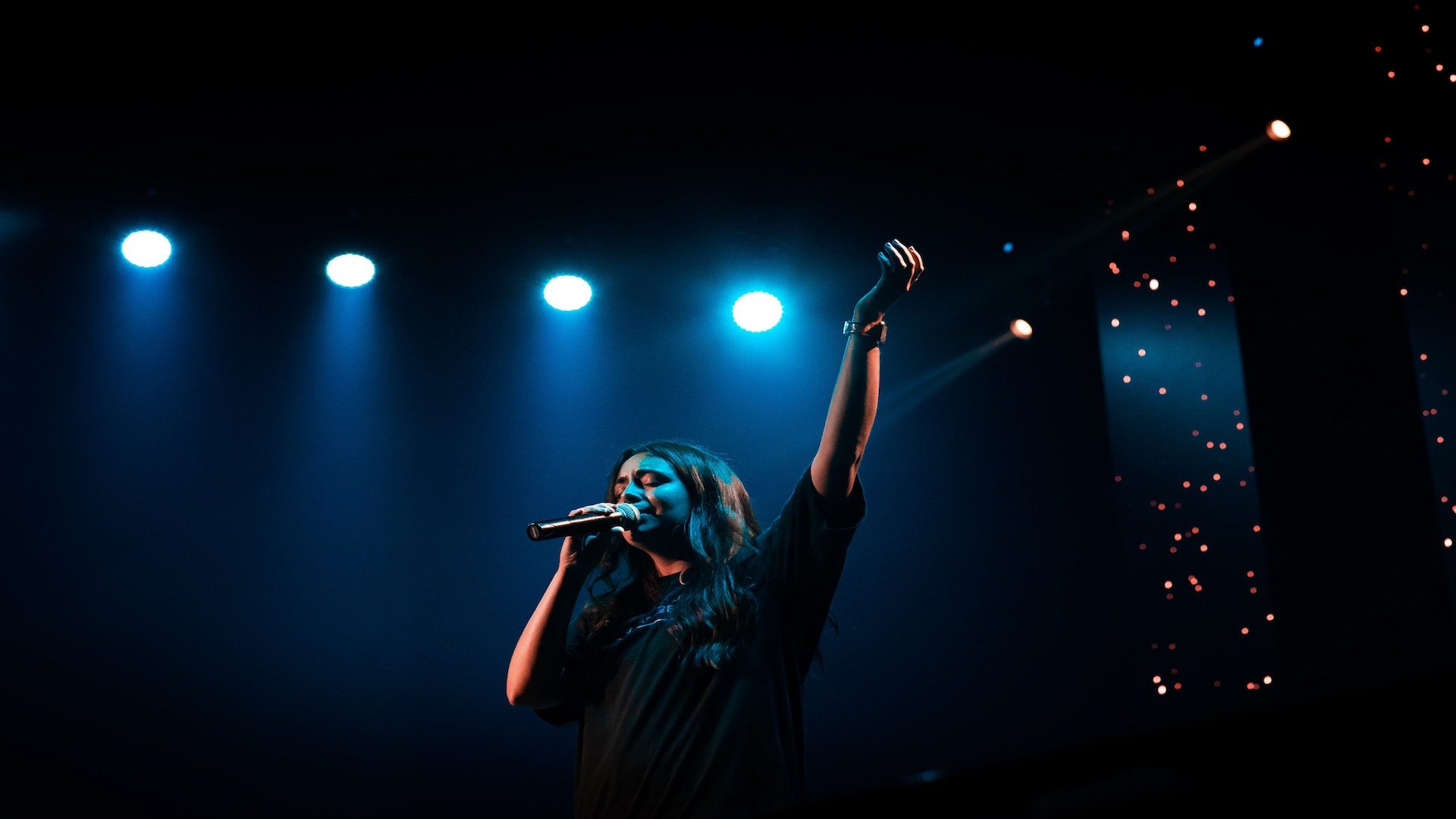A woman in a black shirt singing in a microphone with her arm raised on a stage.