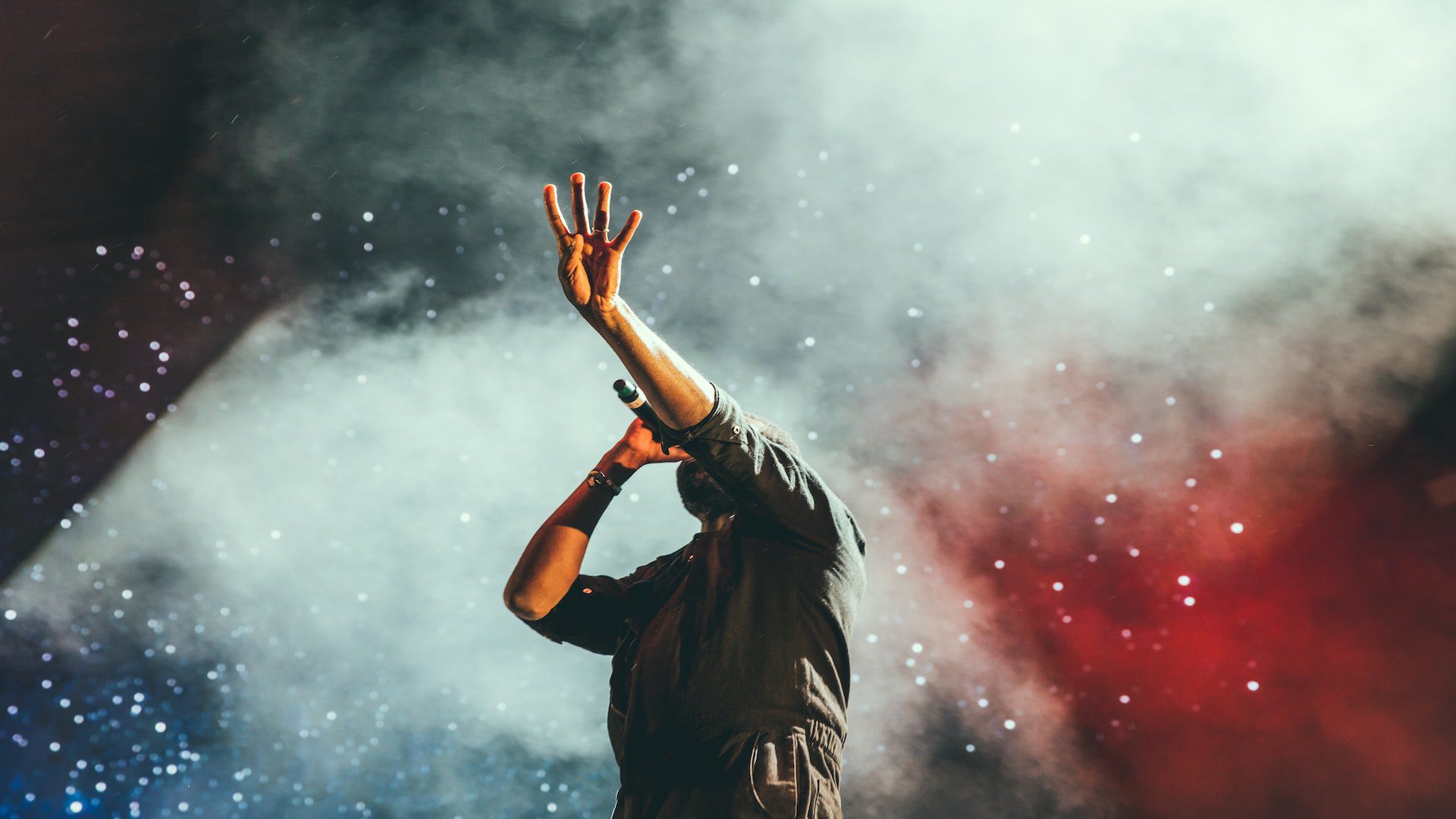 A man with his arm raised singing in a microphone. There are red, blue, and white lights behind him.