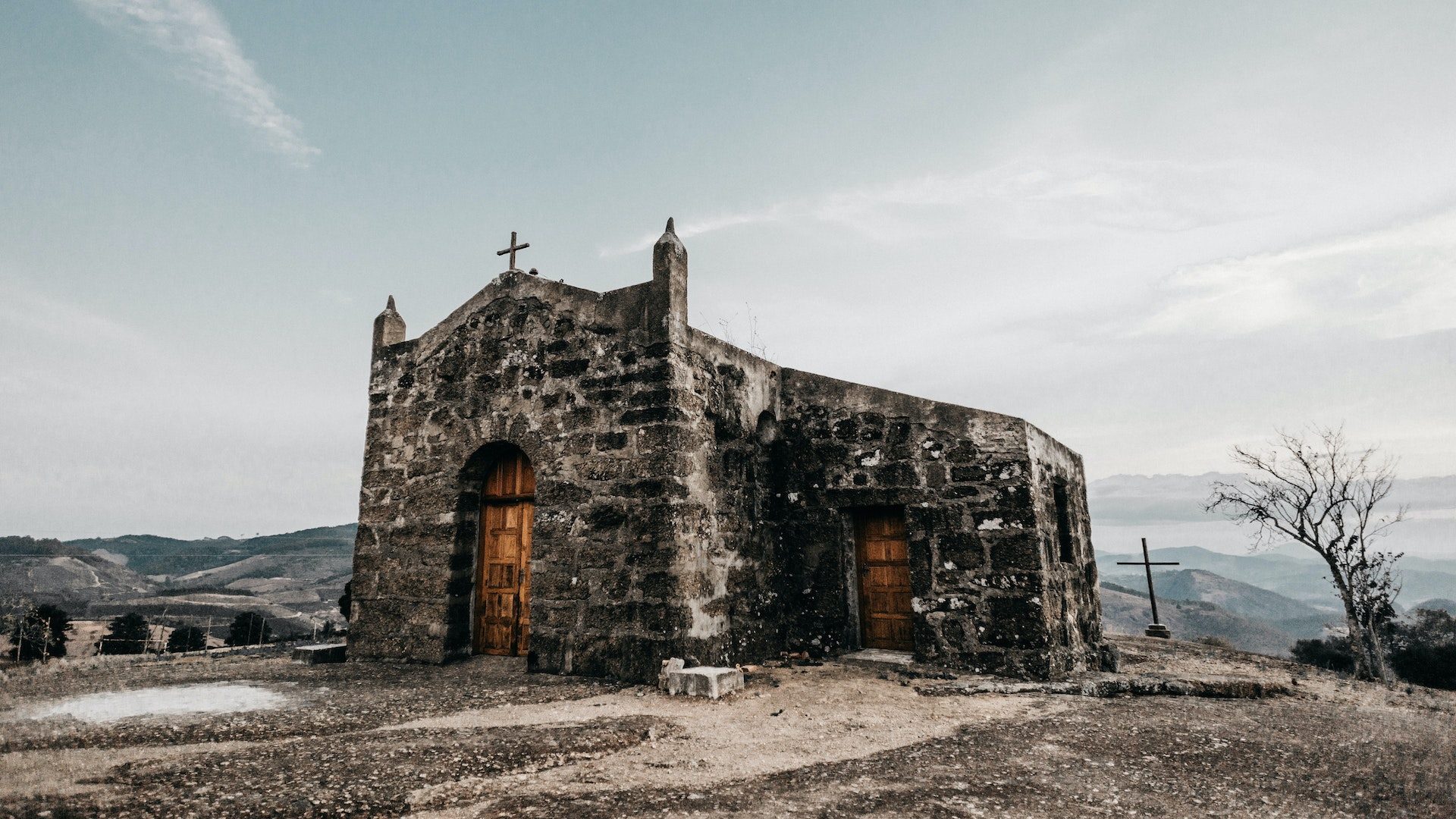 A gray, stone church with wooden doors and a cross on the roof near mountains.