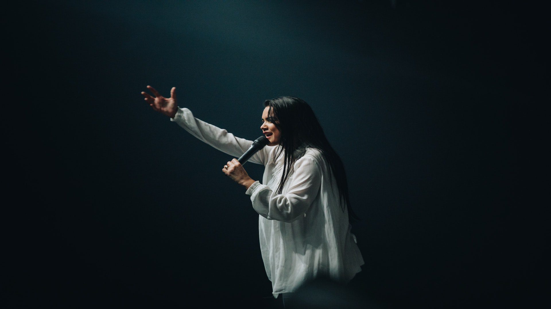 A woman with a white shirt singing in a microphone with her arm raised.