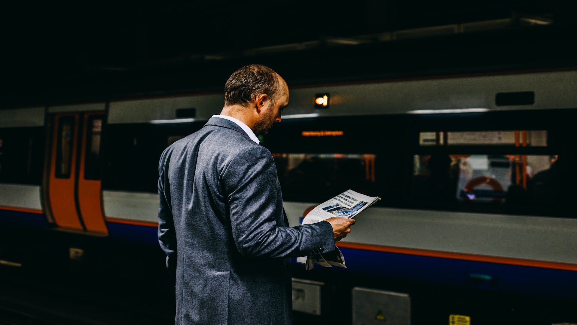 A man in a suit in front of a subway reading a newspaper.