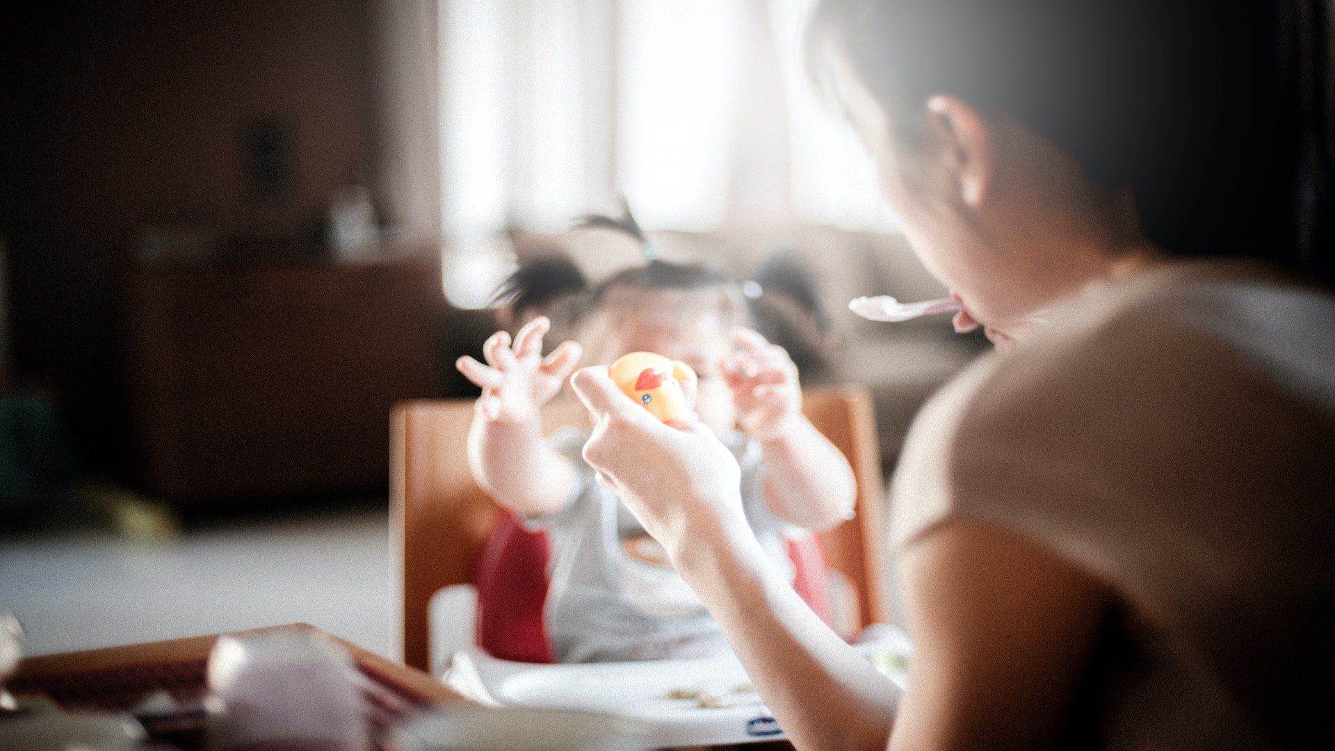 A mother holding a rubber duck while feeding her daughter in a high chair.