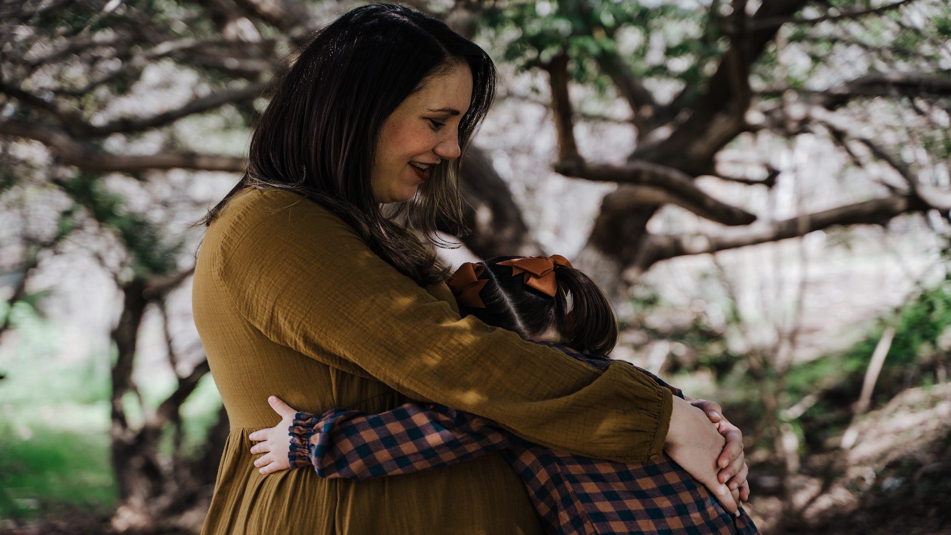 A mother in a yellow shirt hugging her toddler daughter in a plaid dress in front of a tree.