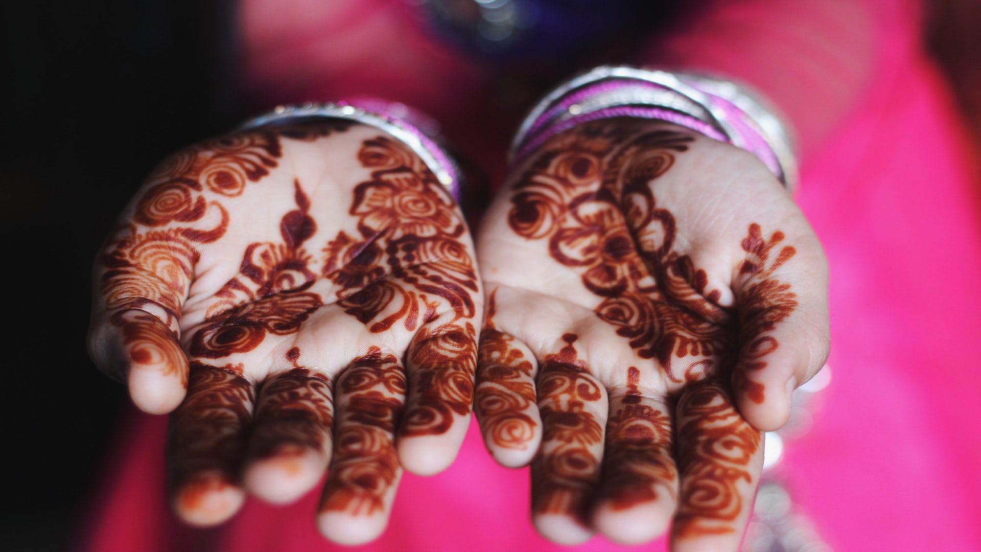 A woman in a pink dress with bracelets holding out her hands with floral henna designs.