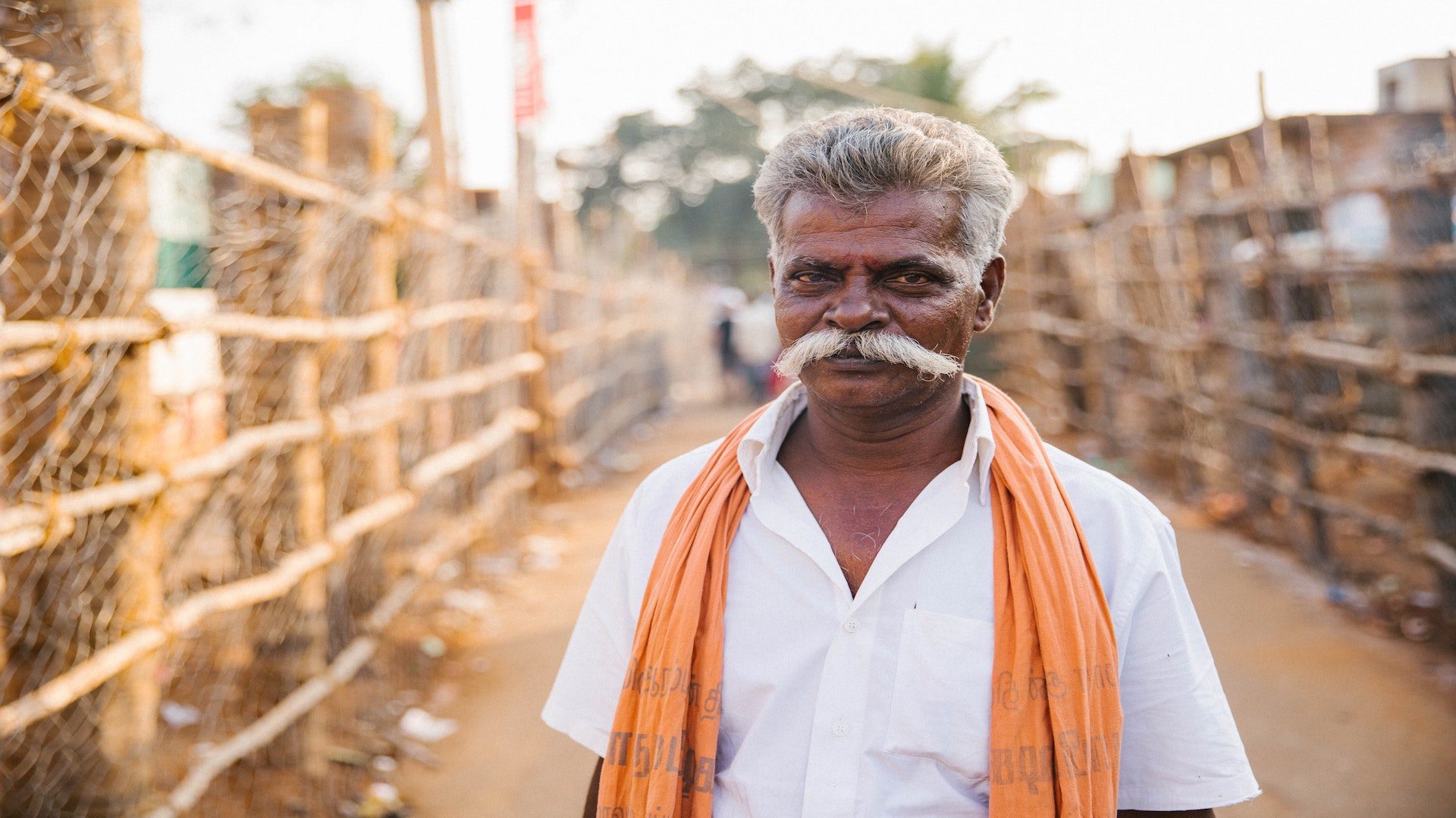 An elderly Indian man with a moustache in a white shirt and orange scarf outside.