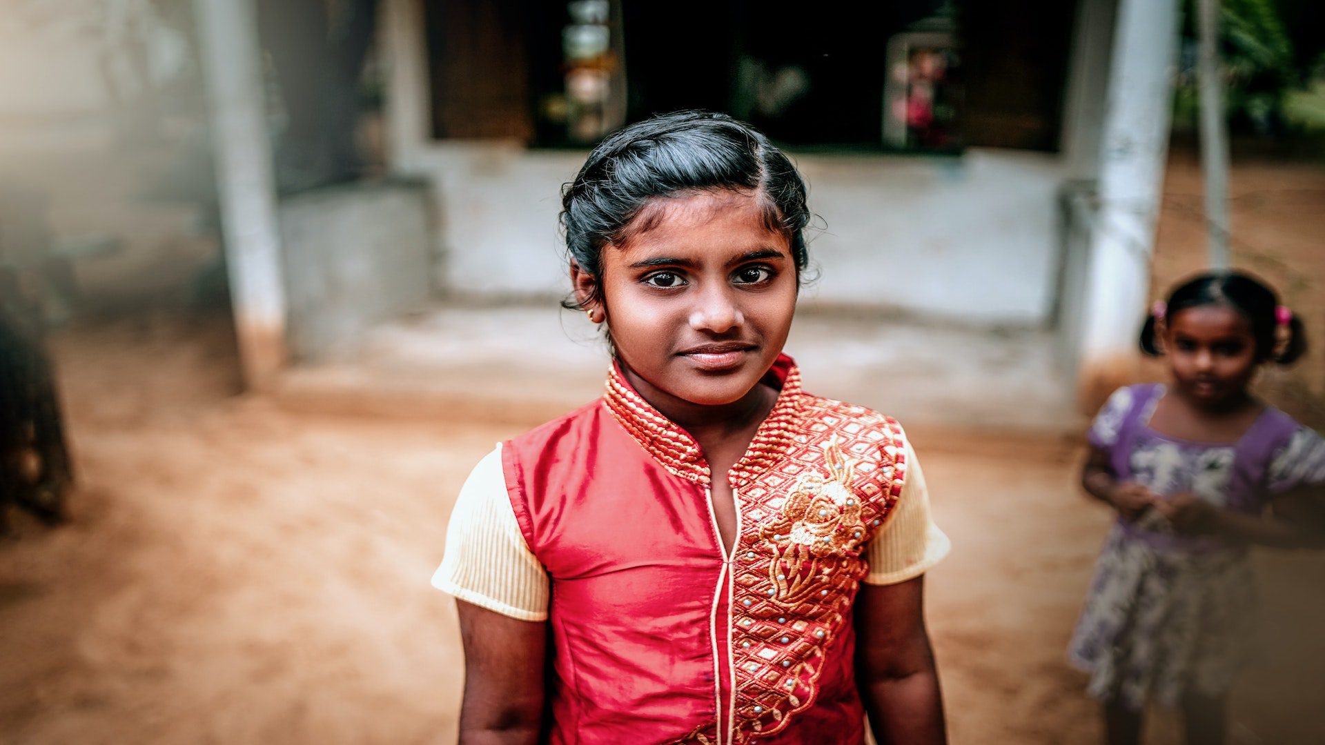 Young indian girl in a red dress standing on dirt in front of a white building.