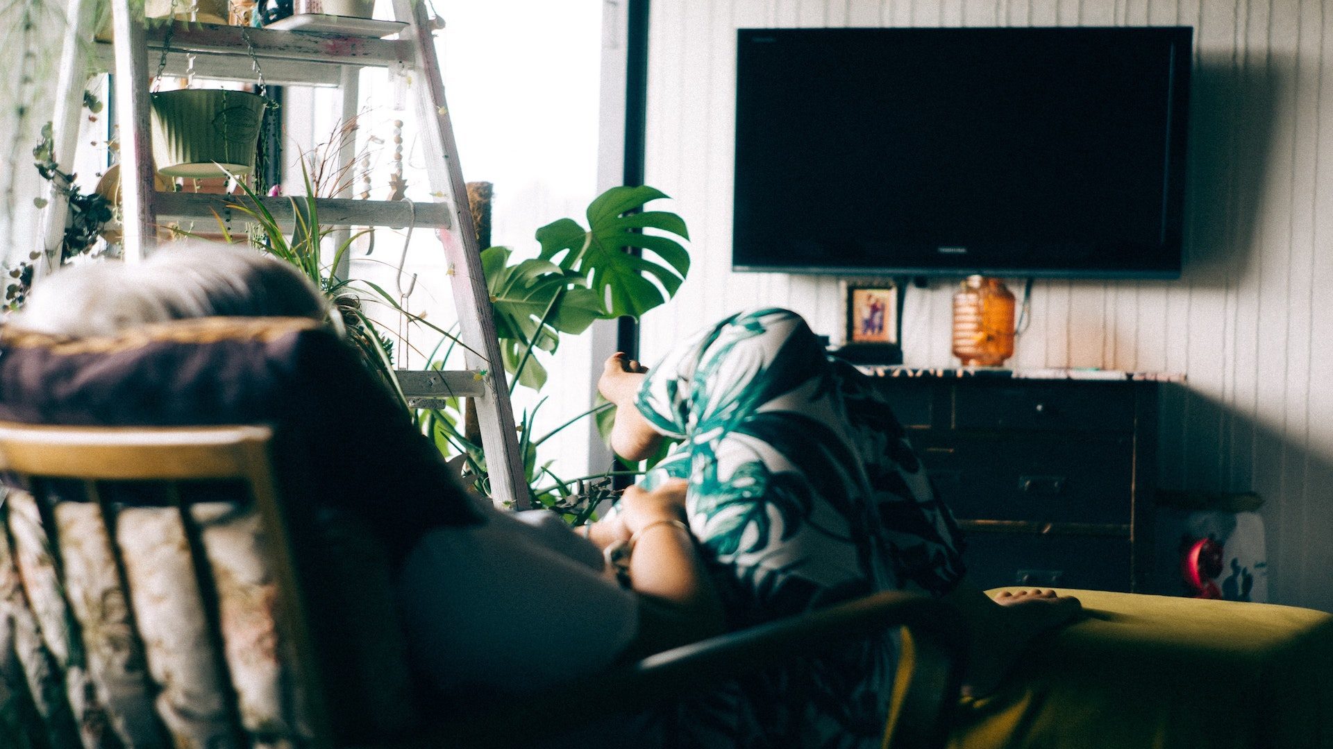 A woman sitting on a chair next to green plants watching a television.