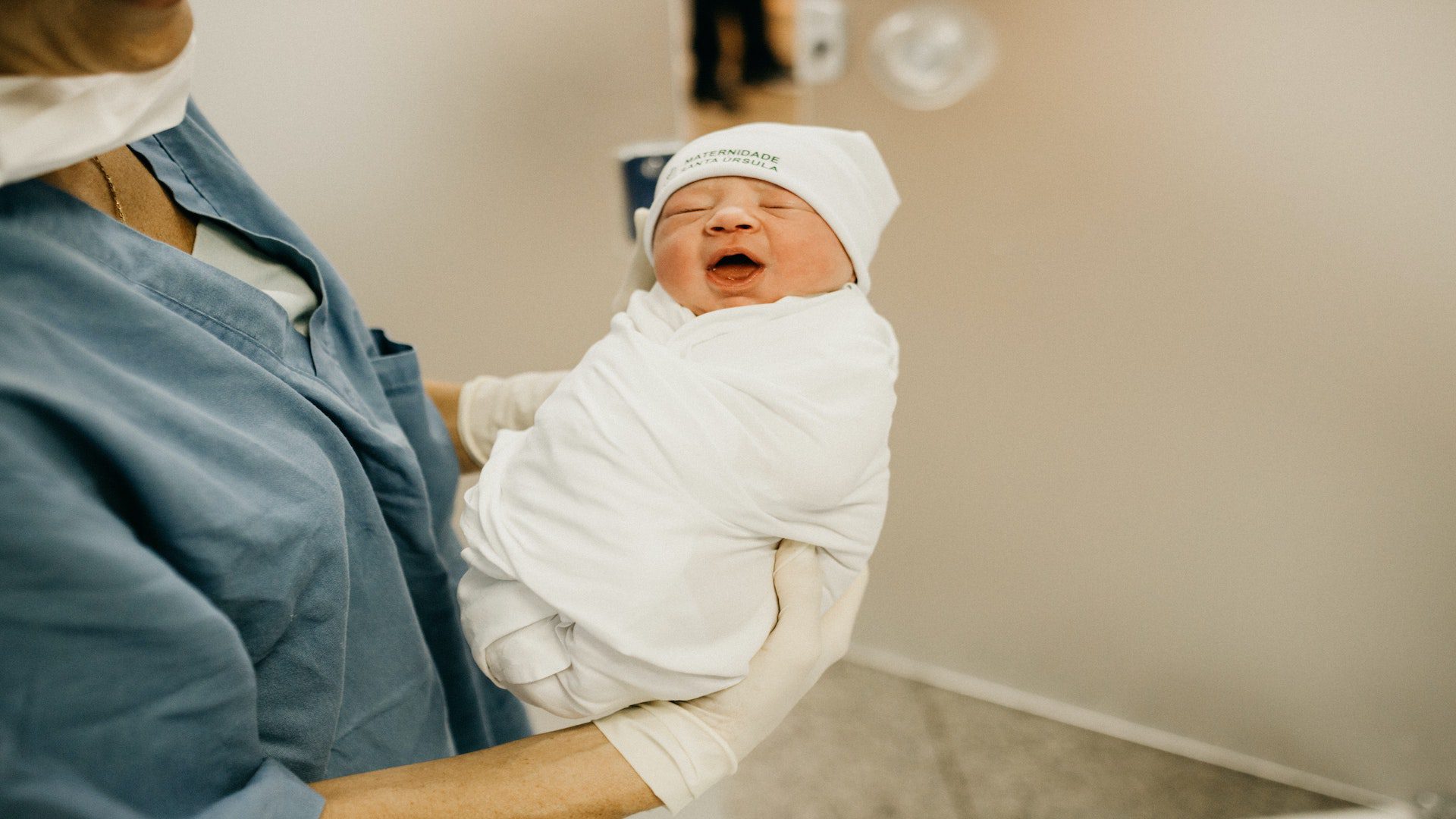 A nurse holding a newborn baby with a white hat wrapped in a white blanket.