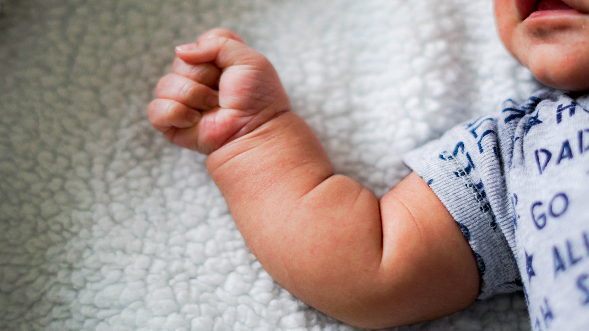A baby lying on a white blanket with its hand in a fist.