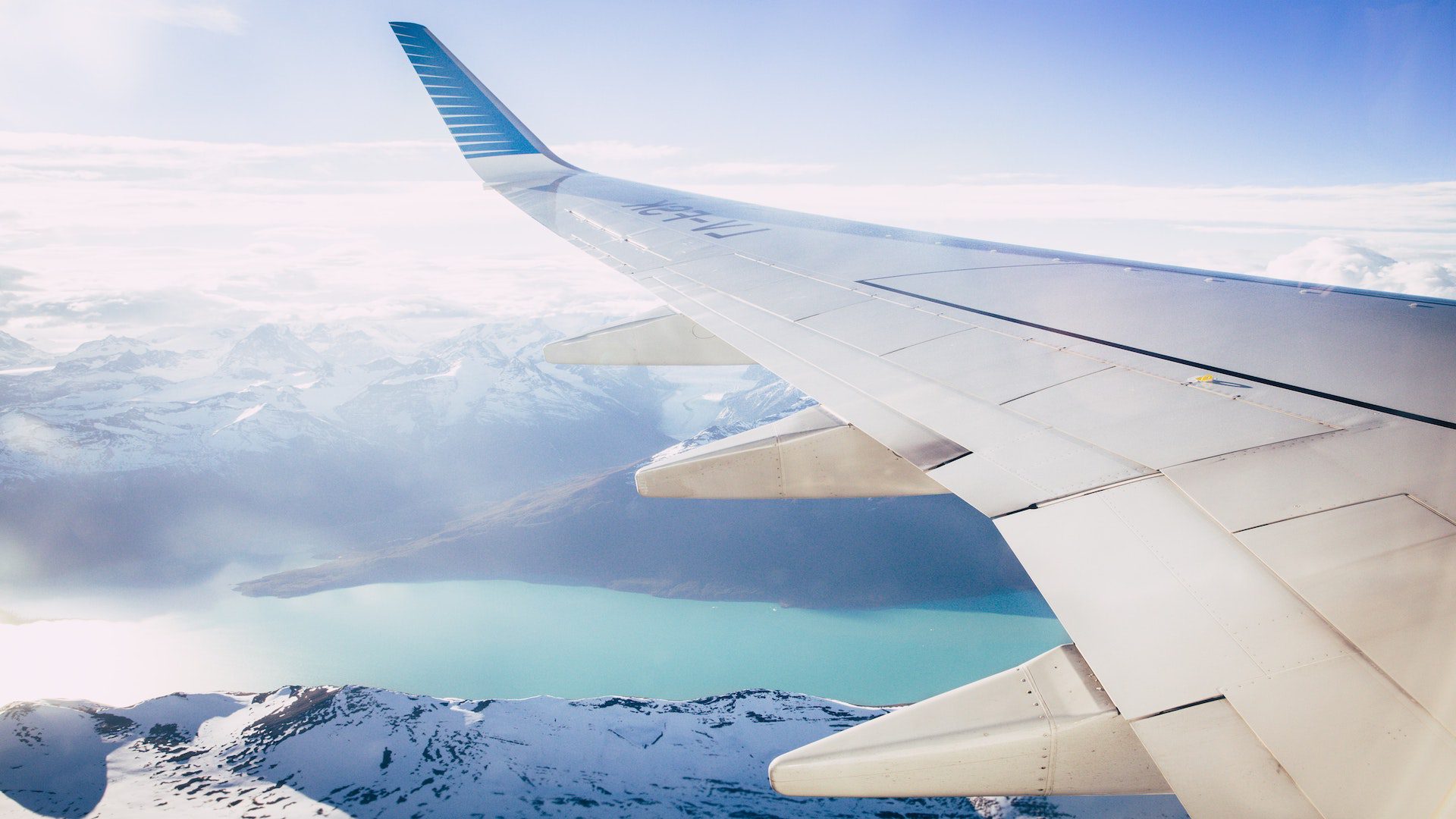 White wing of a plane over snow covered mountains and a river.