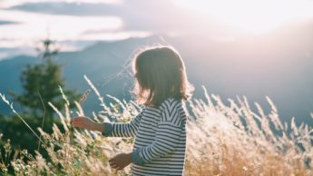 The side of a young girl in a striped shirt standing in front of wheat and mountains.