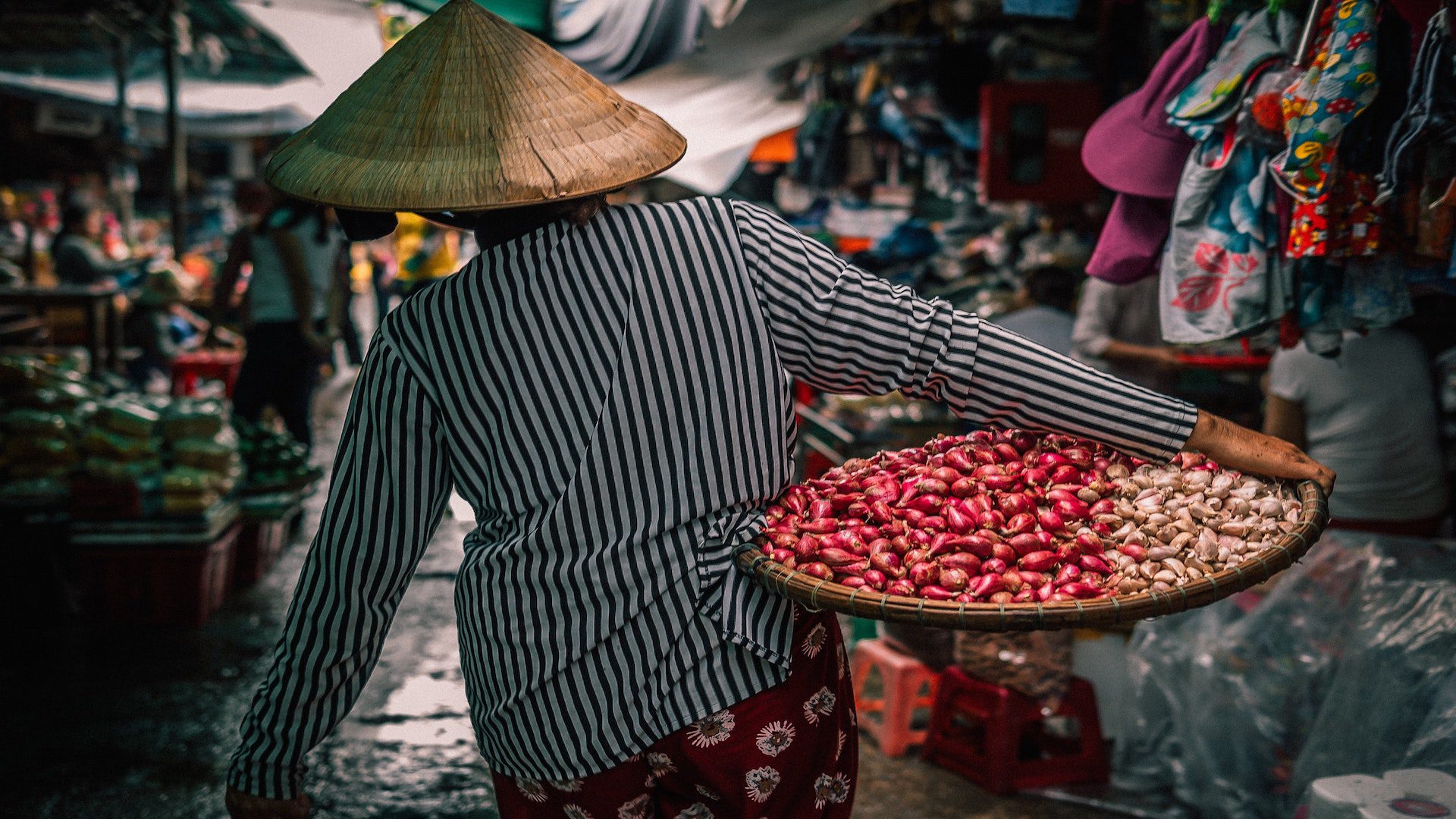 The back of a person in a striped shirt and straw hat on a street carrying a basket with red and white fruit.