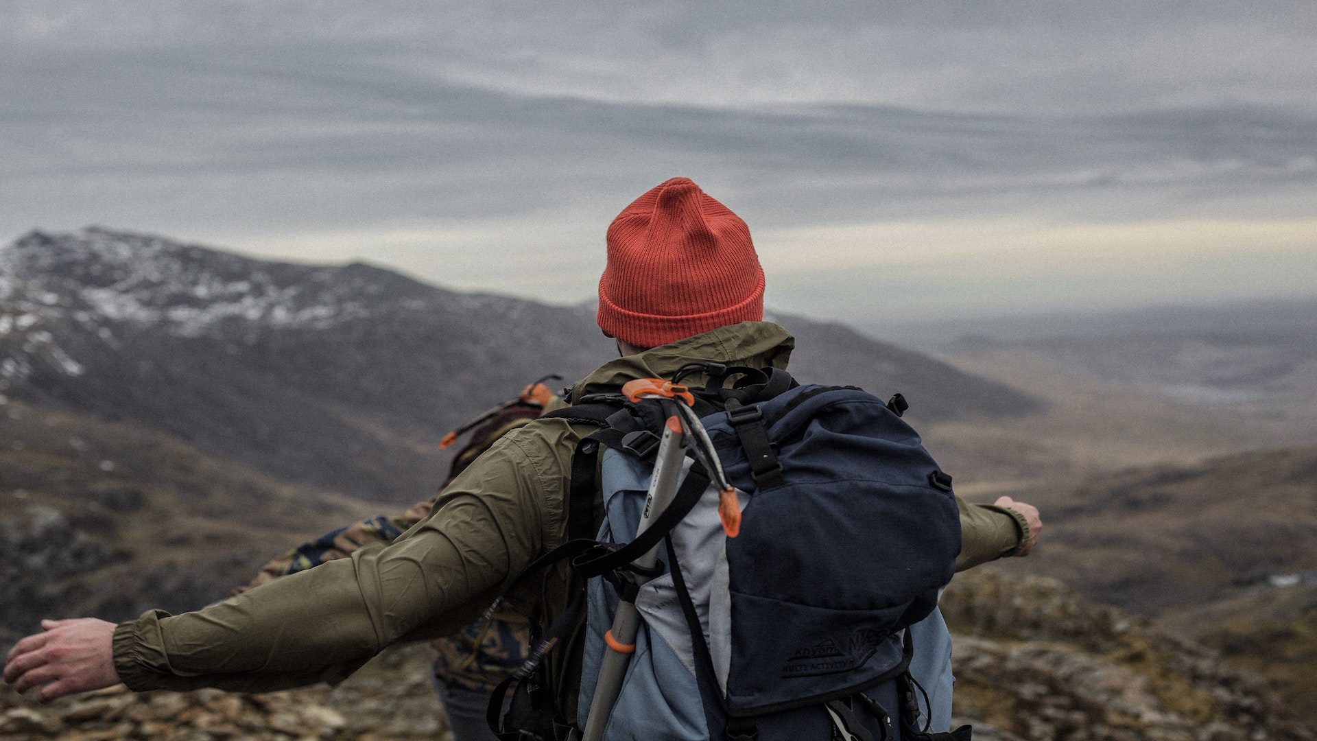 The back of a man with a hiking backpack with his arms outstretched on a mountain.