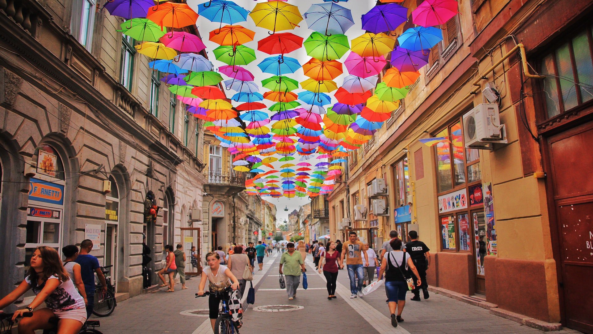 A group of people walking down a street with an asortment of brightly colored umbrellas hanging over the street.