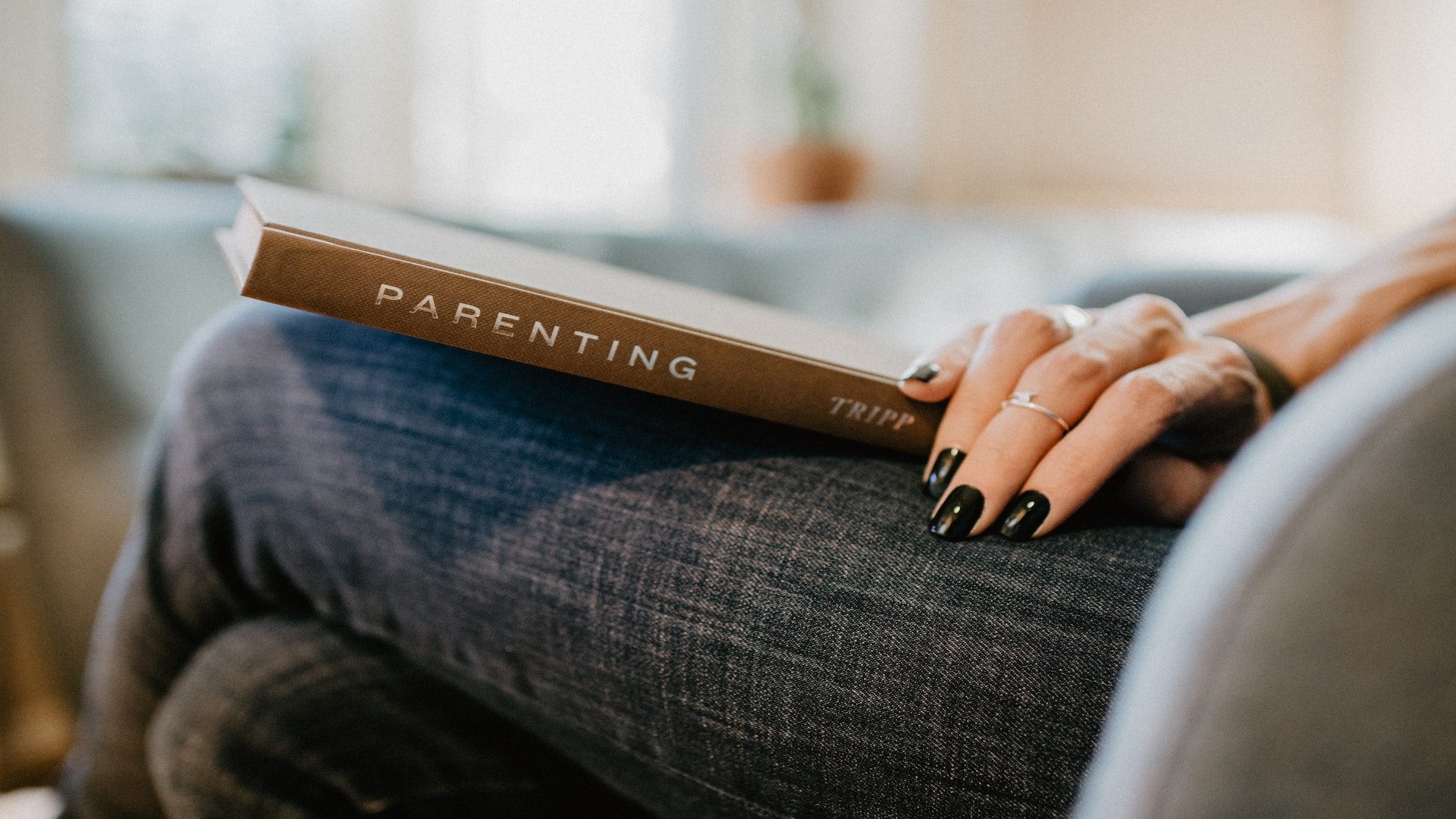 A woman with black nail polish is holding a book titled "Parenting" in her lap.