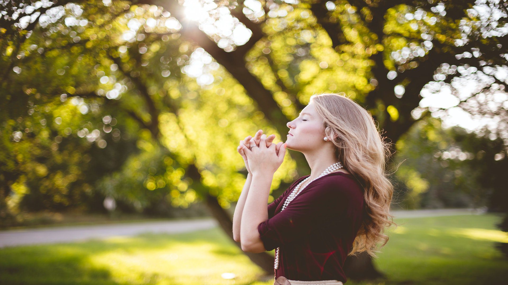 A woman wearing a maroon shirt is praying underneath a tree.