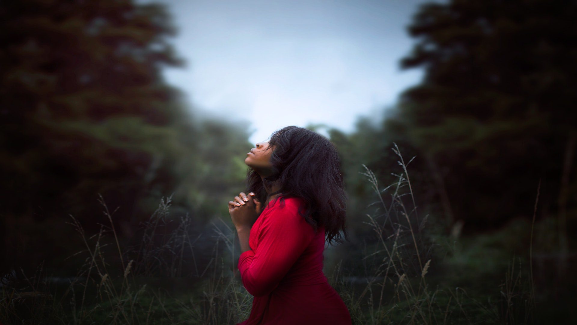 A woman wearing a red sweater is praying with her hands together as she looks at the forest.