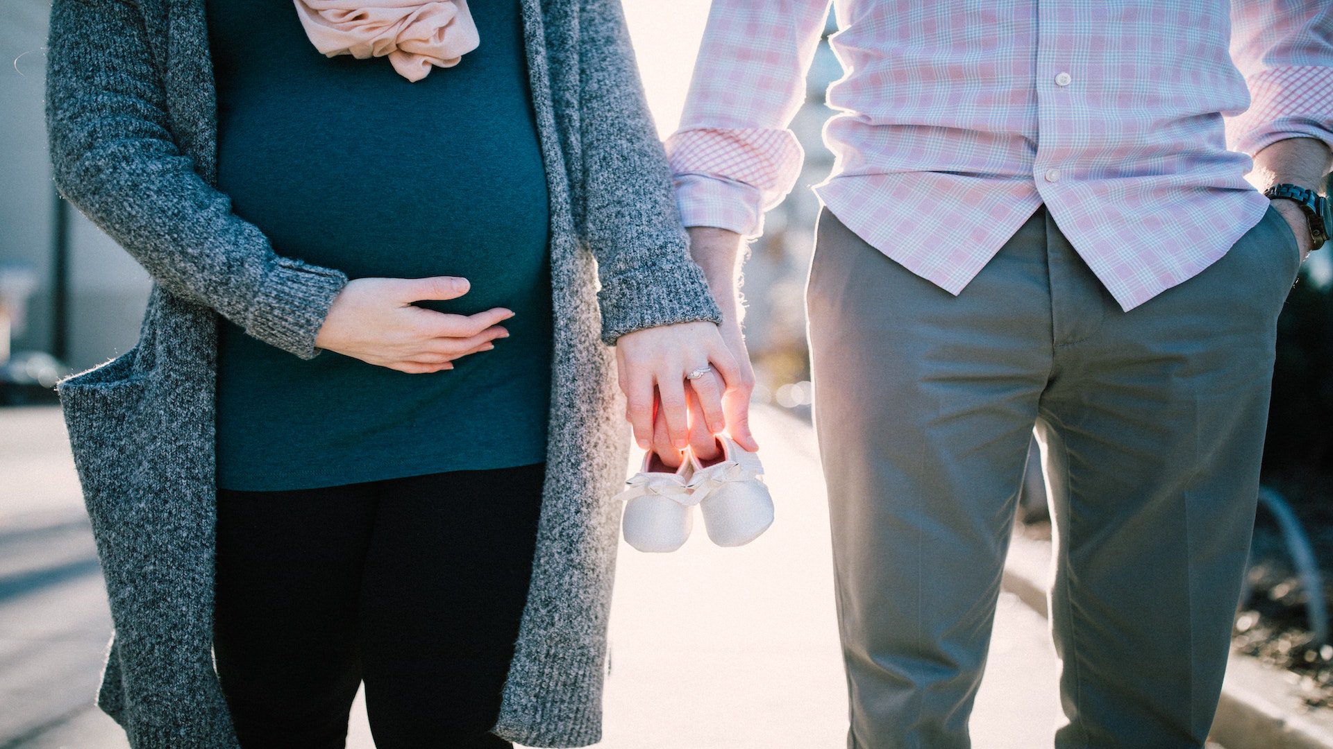 A man is stnading beside a pregnant woman and they are holding the future baby's shoes.