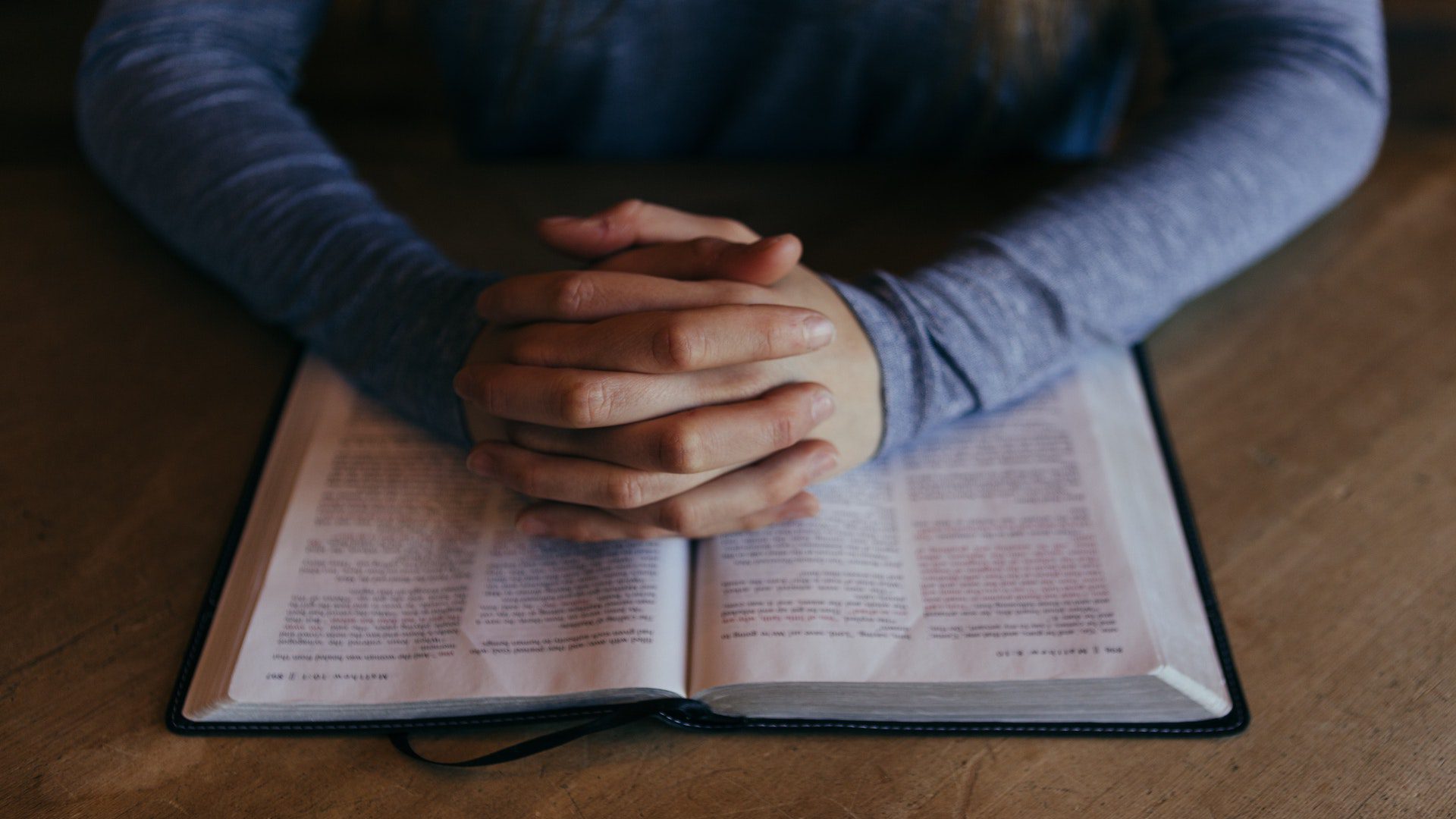 A man wearing a blue long sleeve shirt is holding his hands together over an open Bible.