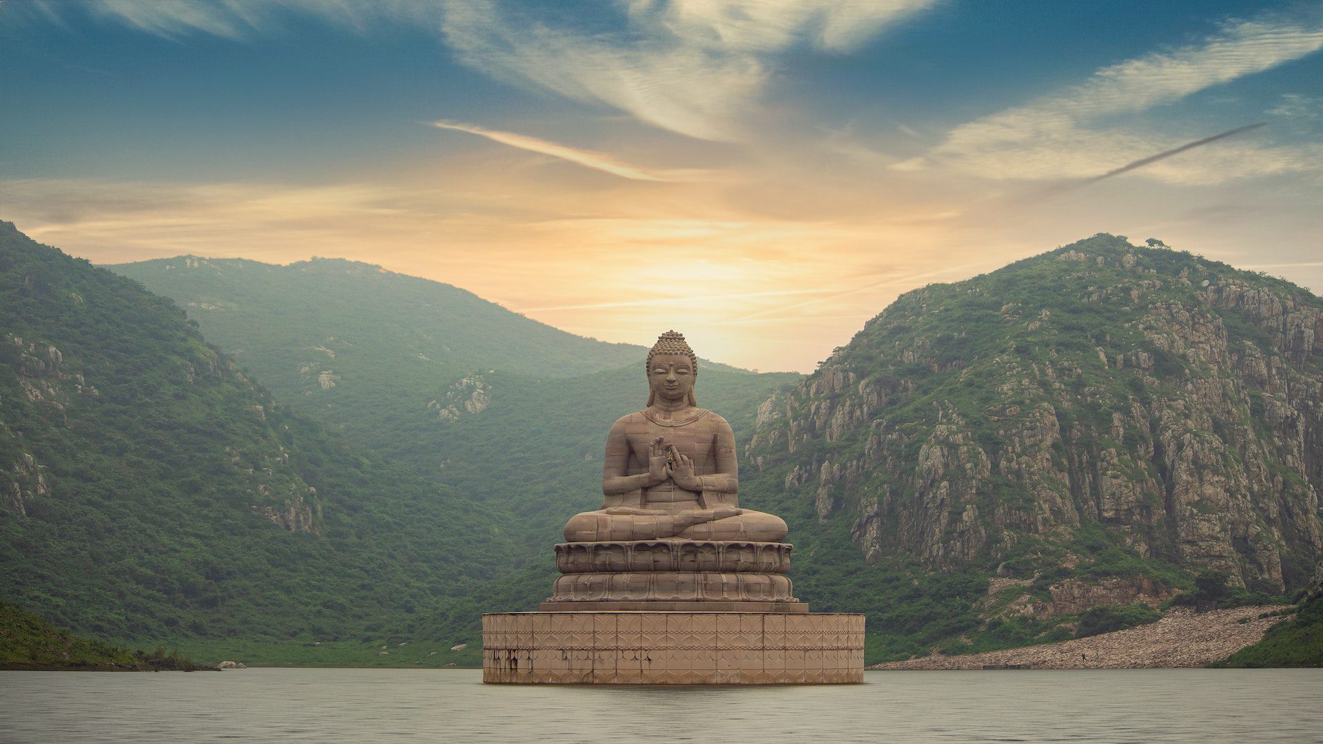 A concrete Buddha statue is floating in a body of water near a green mountain.