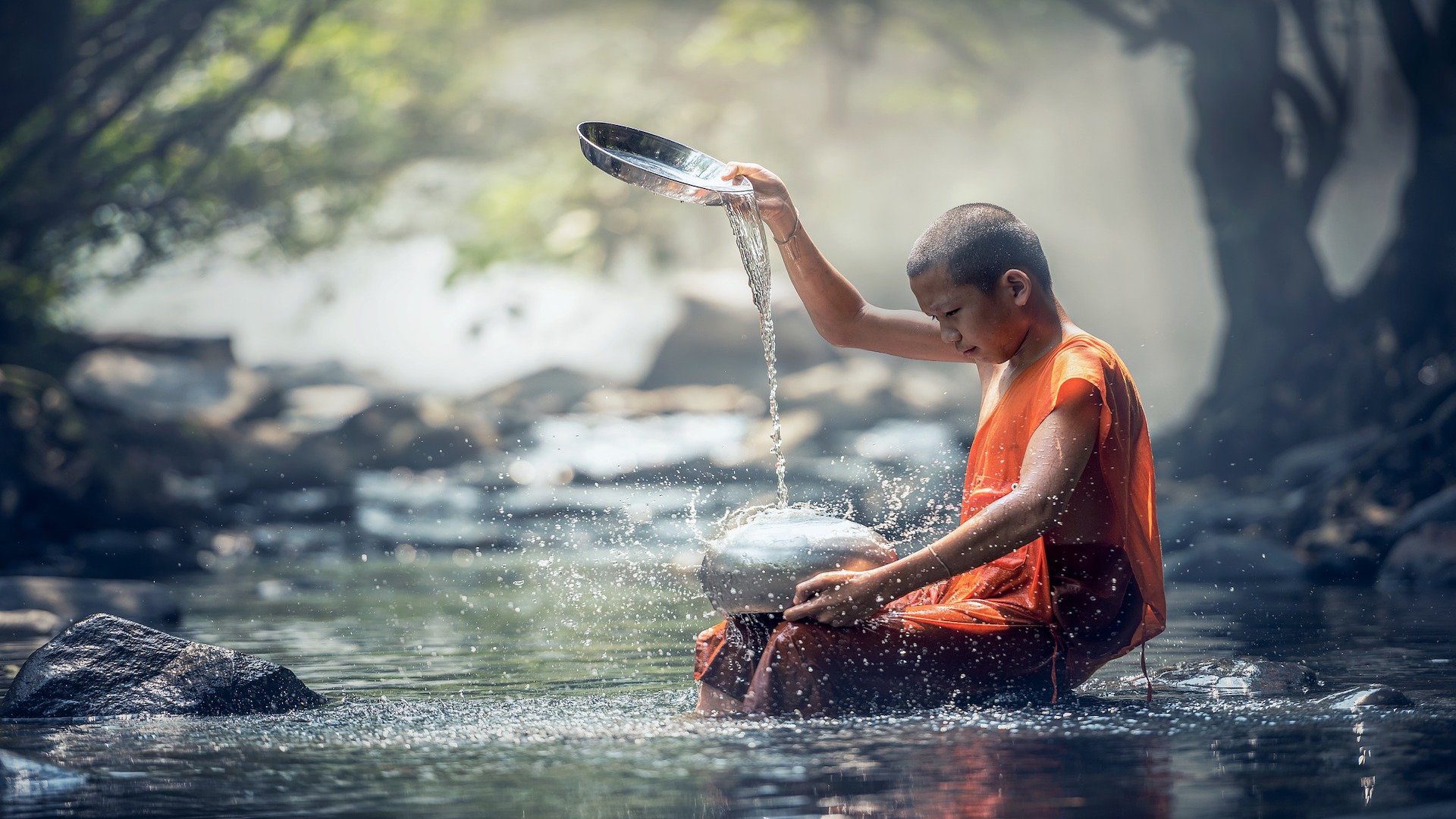 A young male monk is sitting on a rock in a shallow river as he cleans two metal pots.