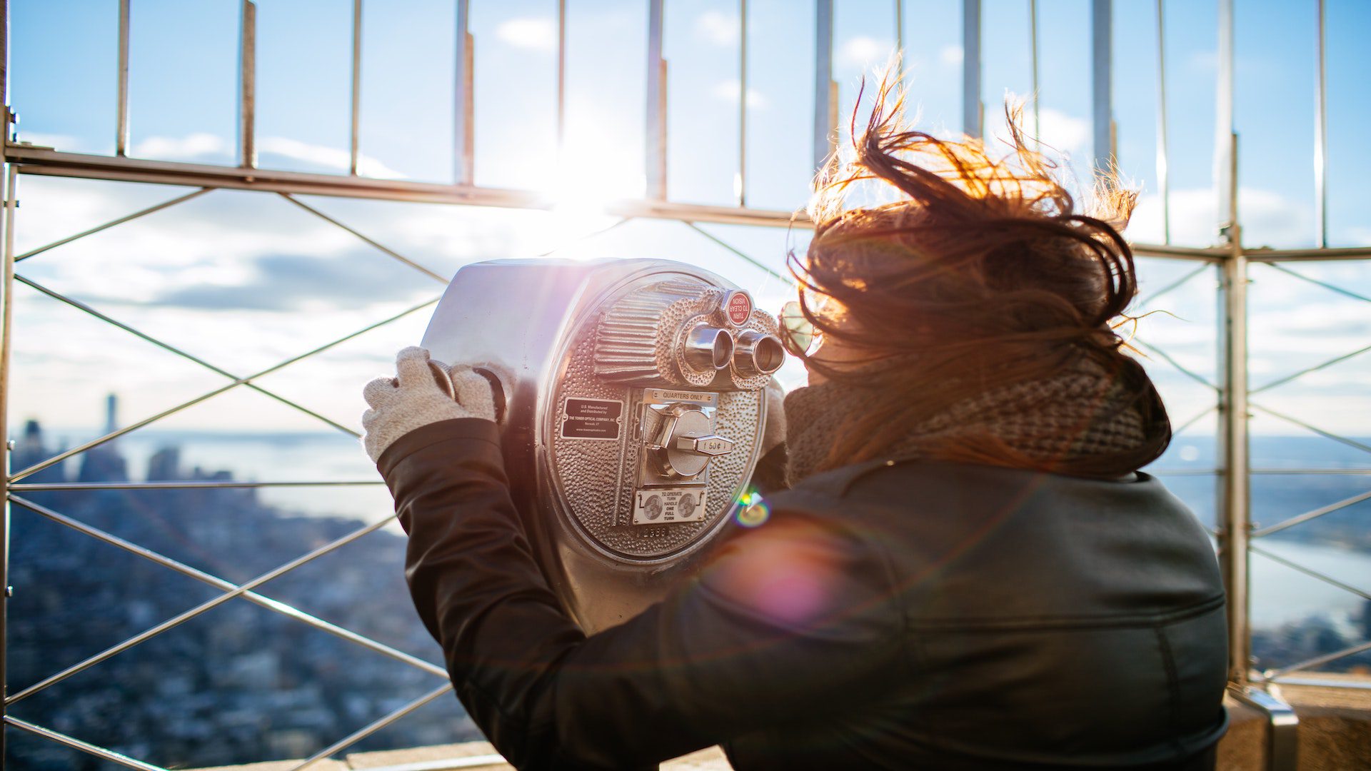 A woman is looking out into the city through a silver viewfinder while on a trip in the city.