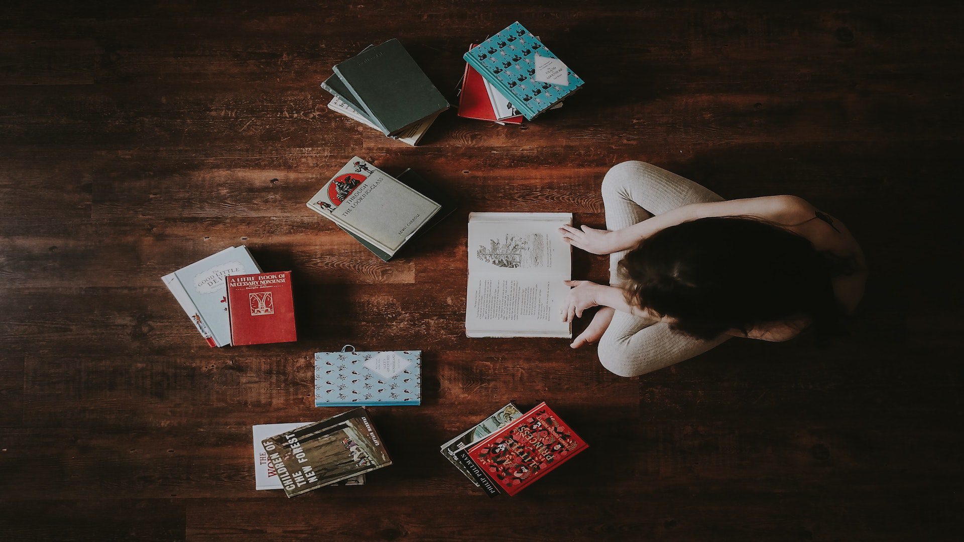 An overhead photo of a woman sitting on a brown wooden floor surrounded by a group of open books.