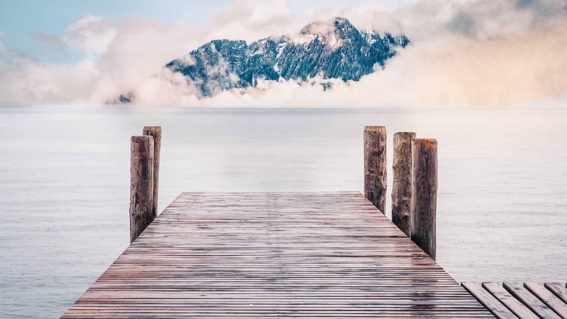 A brown wooden dock that is leading out into a body of water in front of a mountin range looming with white clouds.