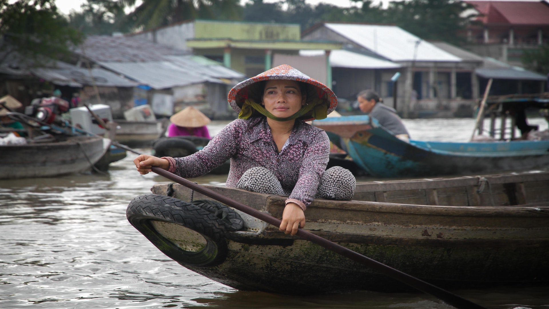 A woman wearing a traditional Vietnamese hat sits at the front of a wooden boat in the water.