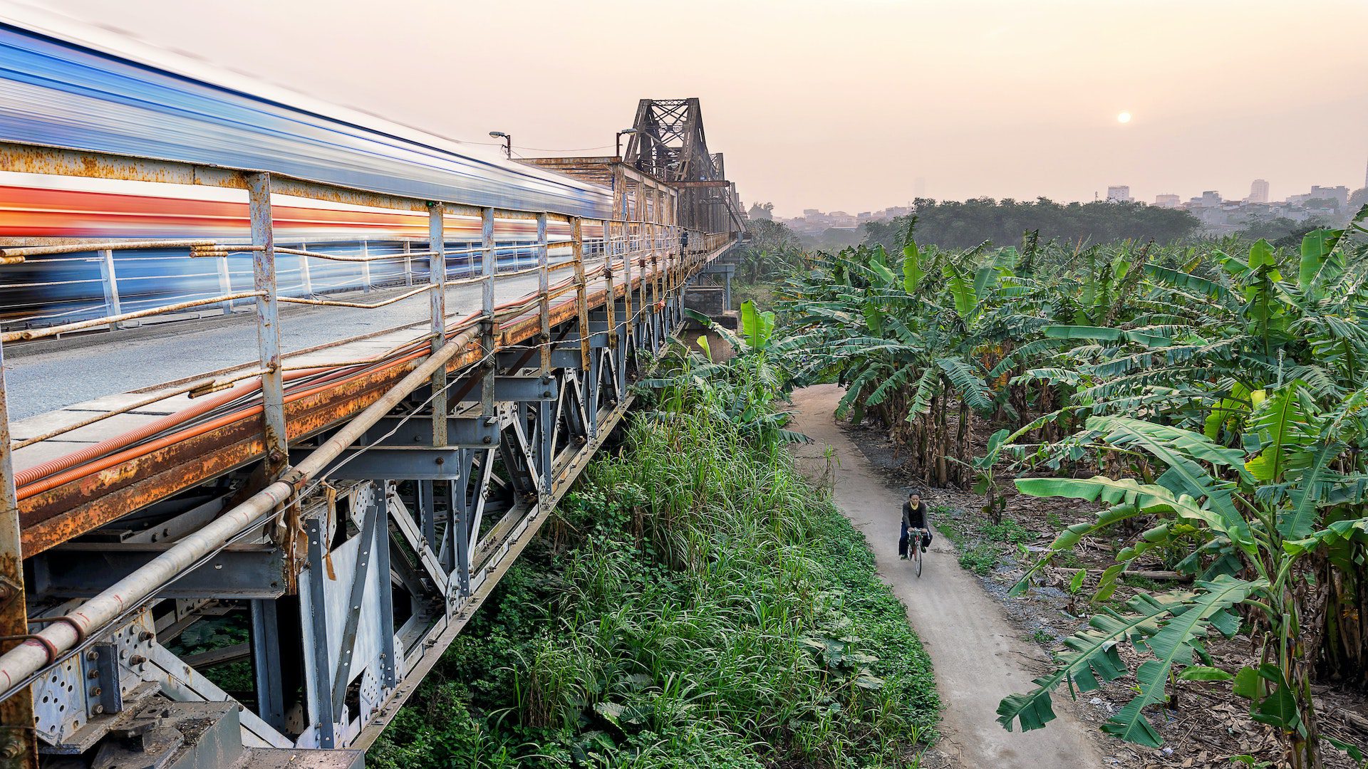 A long exposure photo of a train zooming by on the left and a person biking on a dirt trail on the right.