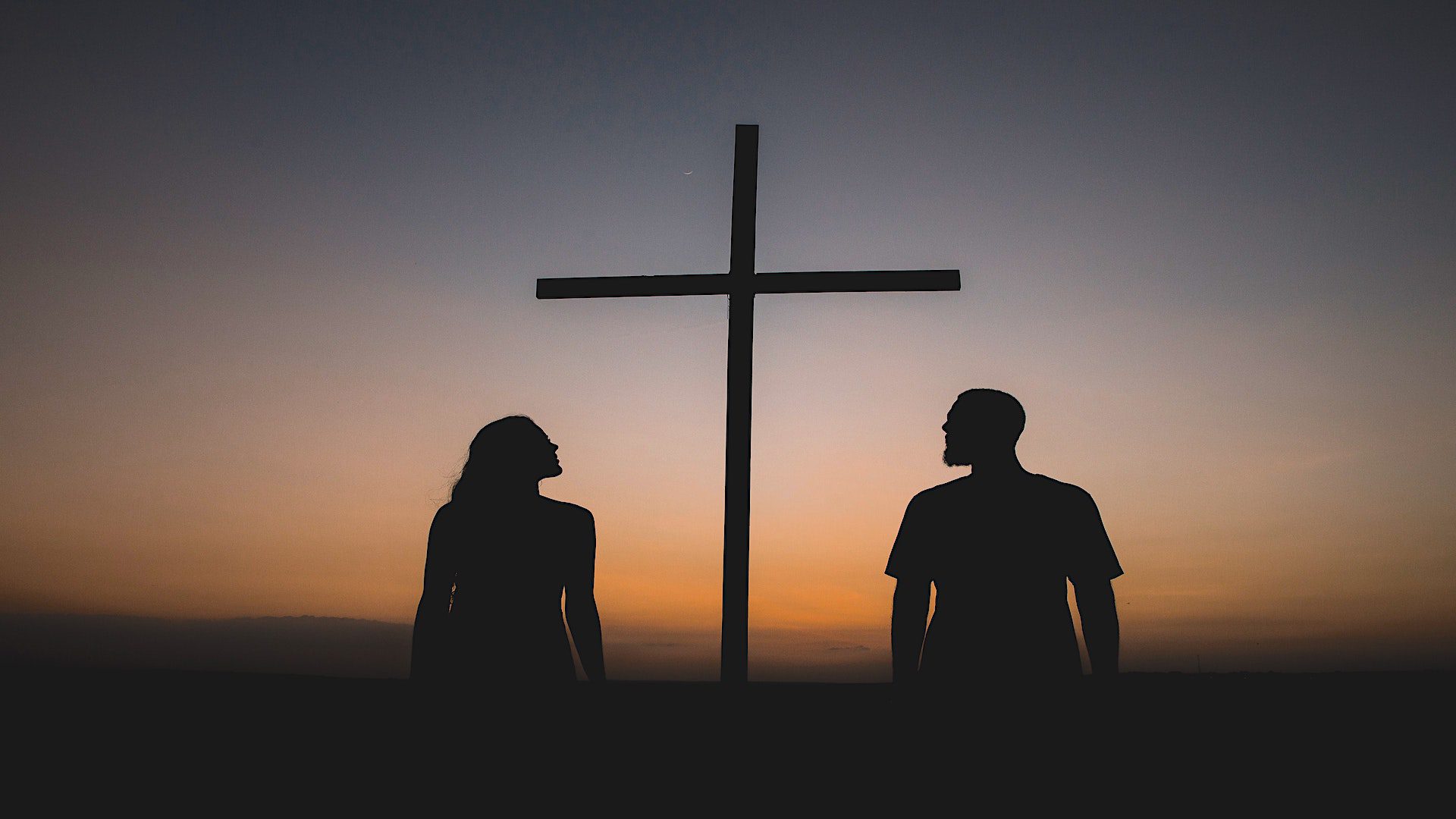 Silhouette of a man and a woman standign beside a wooden cross at dusk.