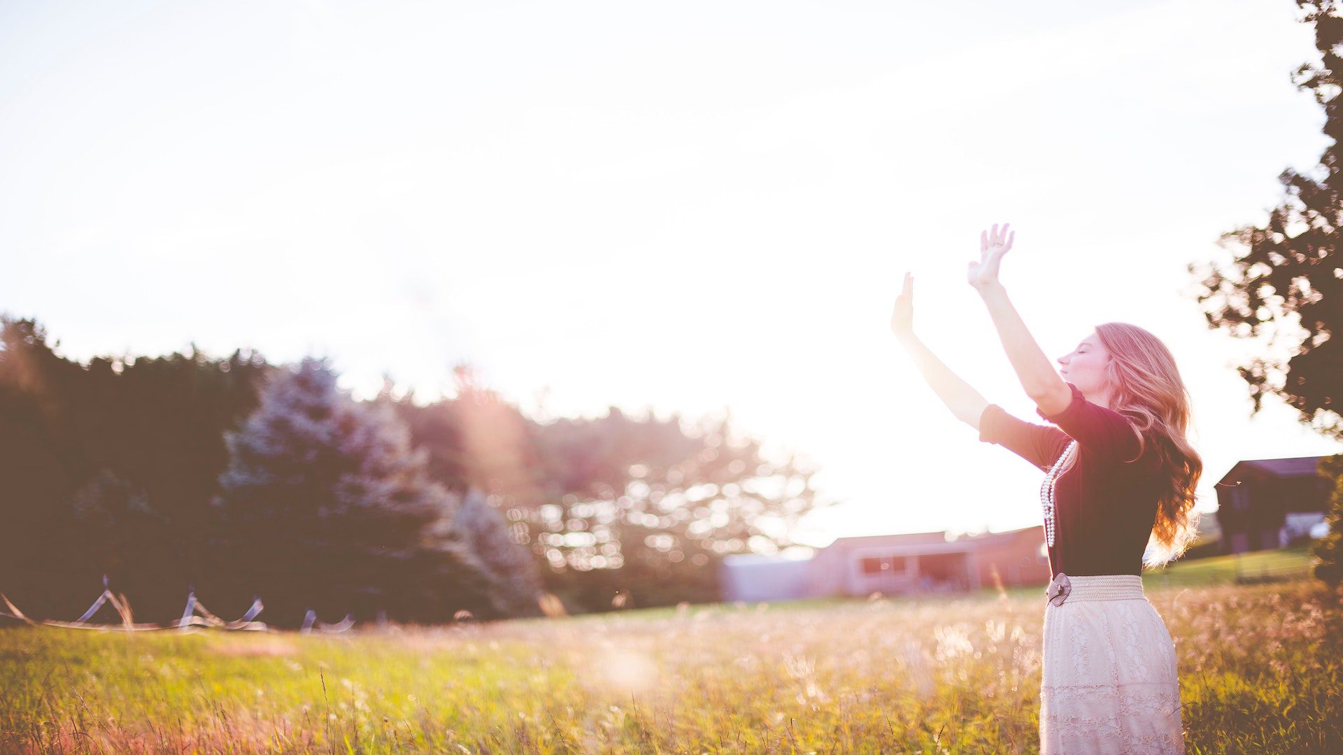 A woman wearing a red top holds her hands up in front of a green meadow with trees.