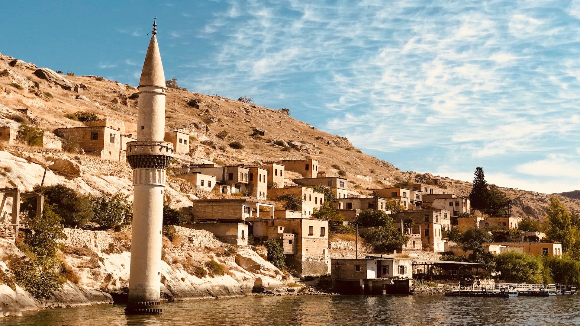 A small rural cluster of buildings and a single tower stnds on the side of a mountain next to a body of water.