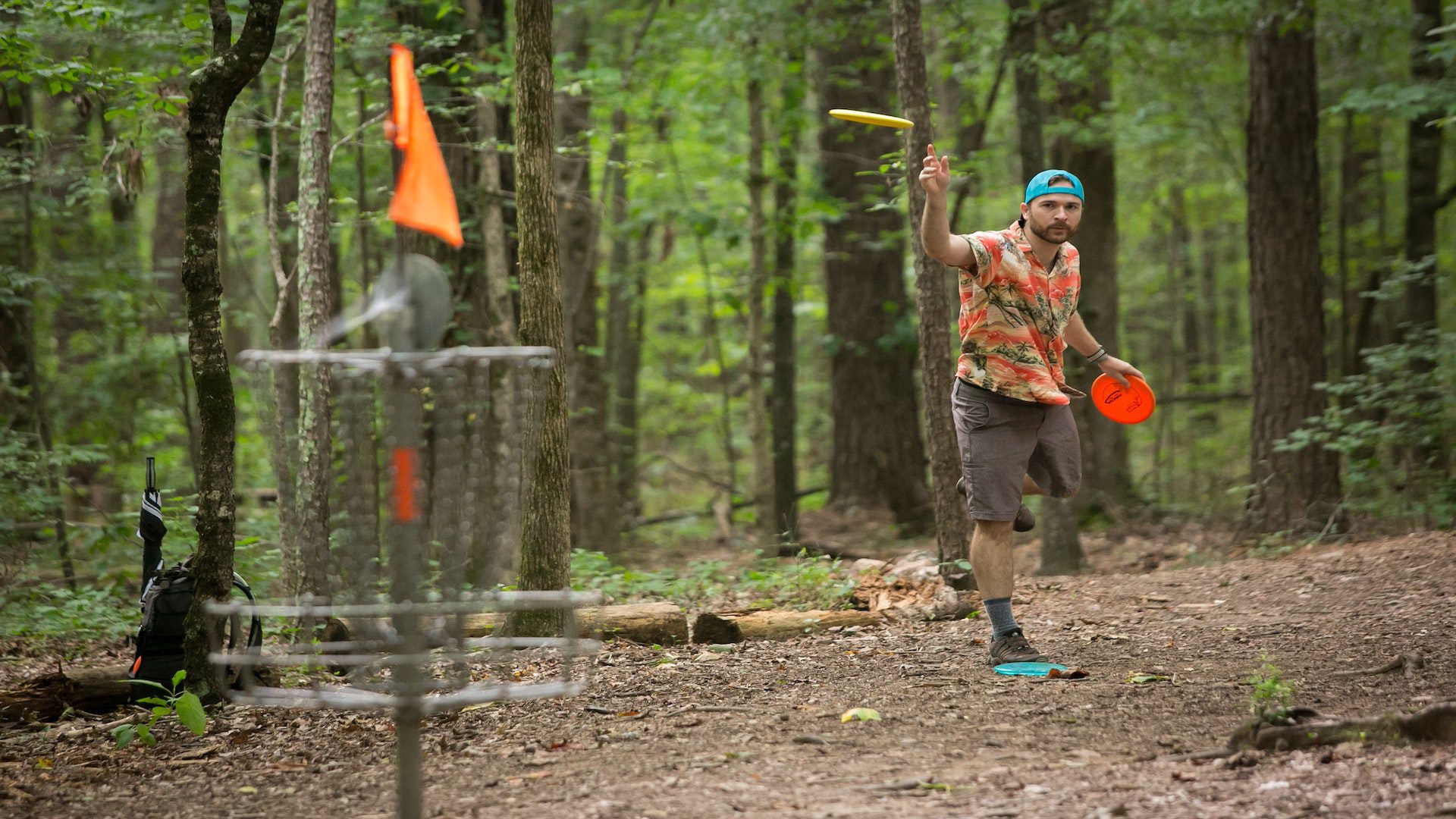 A man in a Hawaiian shirt and baseball cap is throwing a frisbee at a frisbee golf station.