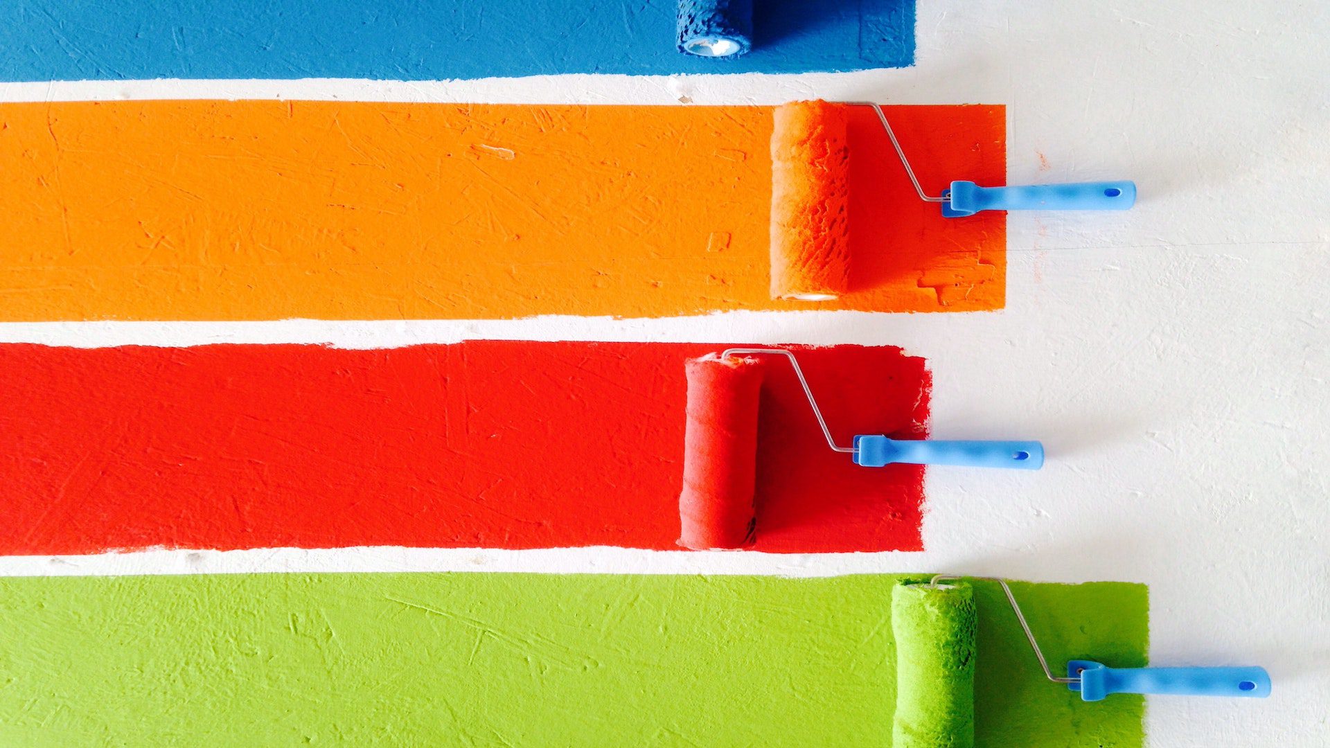 Four paint rollers in the colors of orange, blue, green, and red are painting on a white wall.