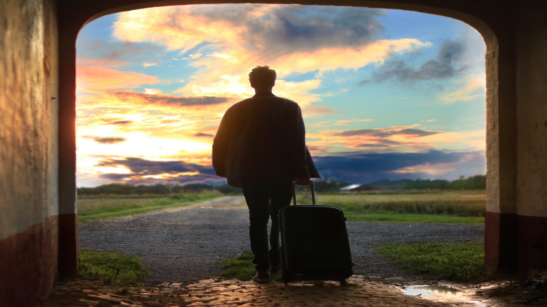 The silhouette of a man holding rolling luggage as he walks through a tunnel towards a beautiful sunset.