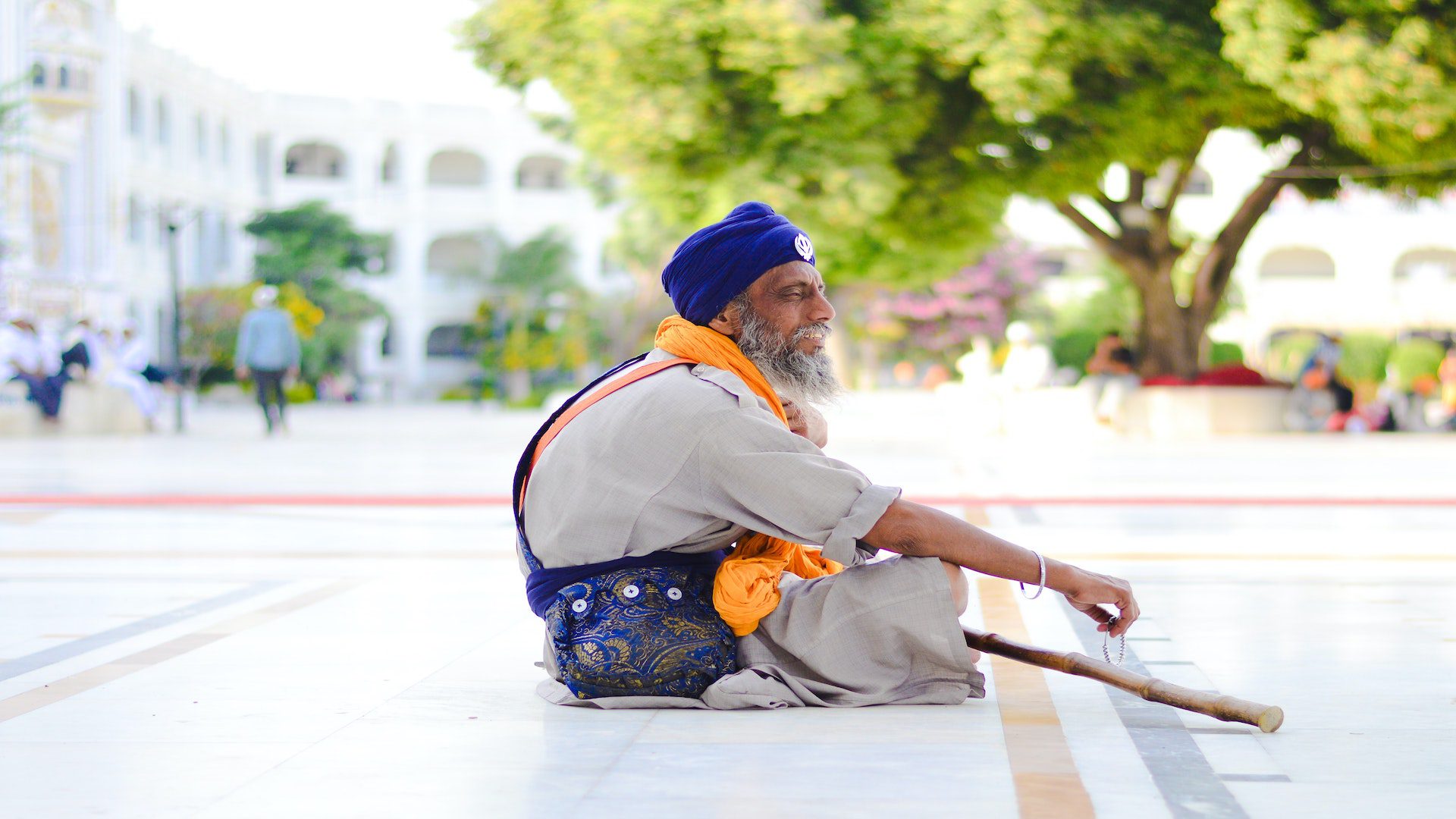 A man is sitting on the gorund with his legs crossed wearing a blue head scarf and orange neck scarf.