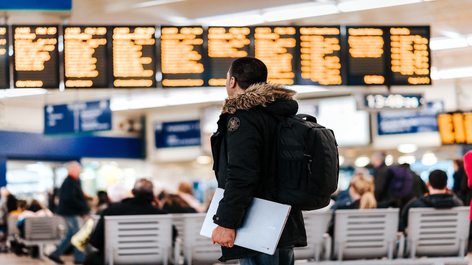A man wearing a black jacket is standing in an airport looking at the arrival/departure board.