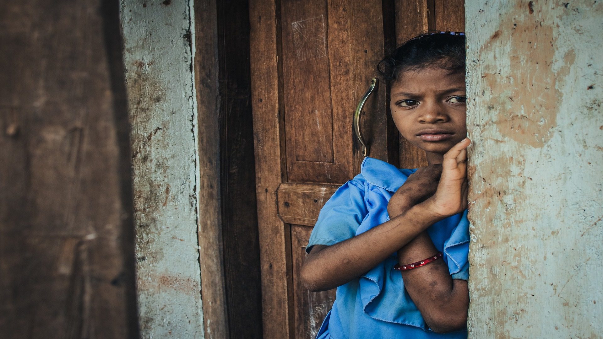 A girl in India is wearing a blue shirt and timidly standing in a doorway.