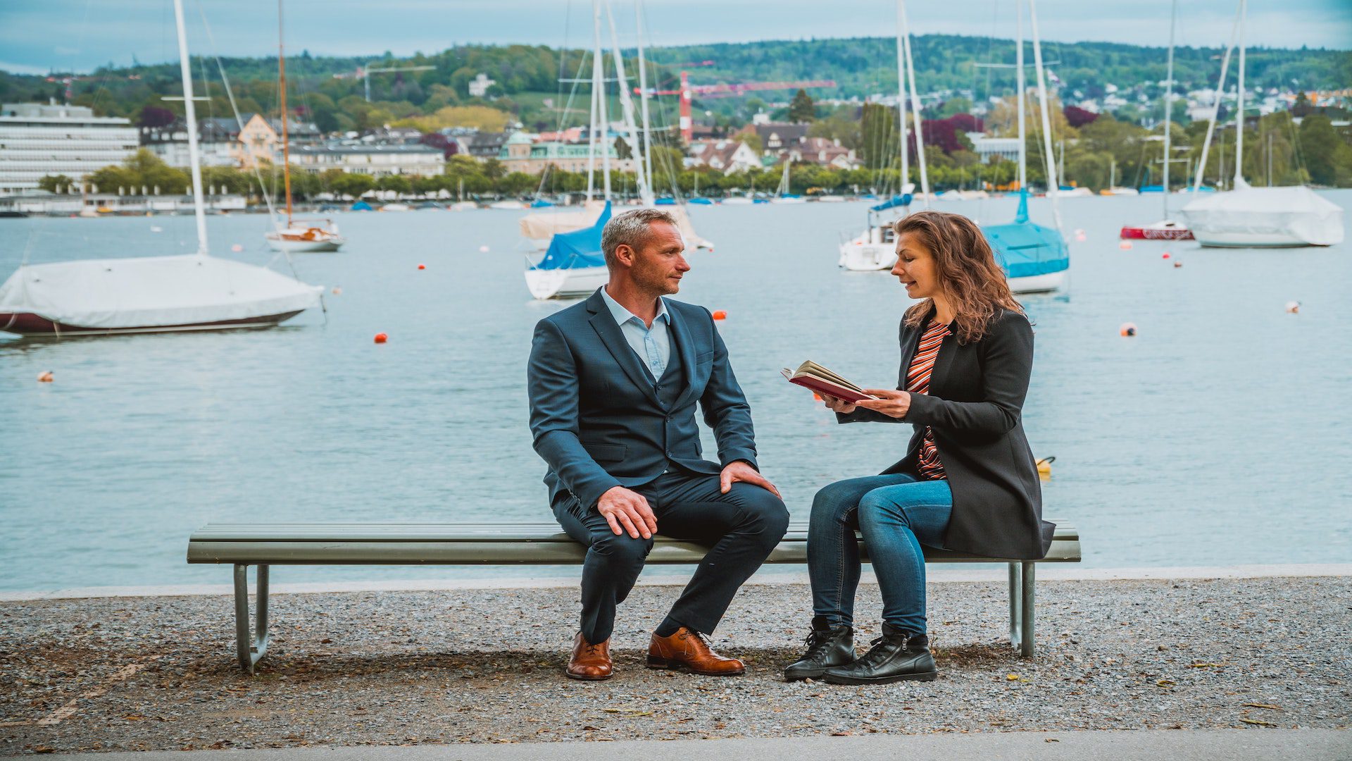 A man and a woman sitting on a bench with sailboats behind them.