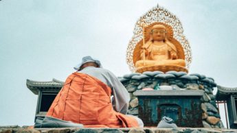 A man in an orange jacket sitting in front of a buddhist statue.