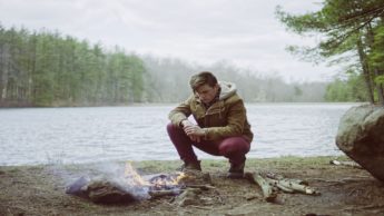 A man in an olive green jacket is crouching down next to the edge of a lake as he examines his camp fire.